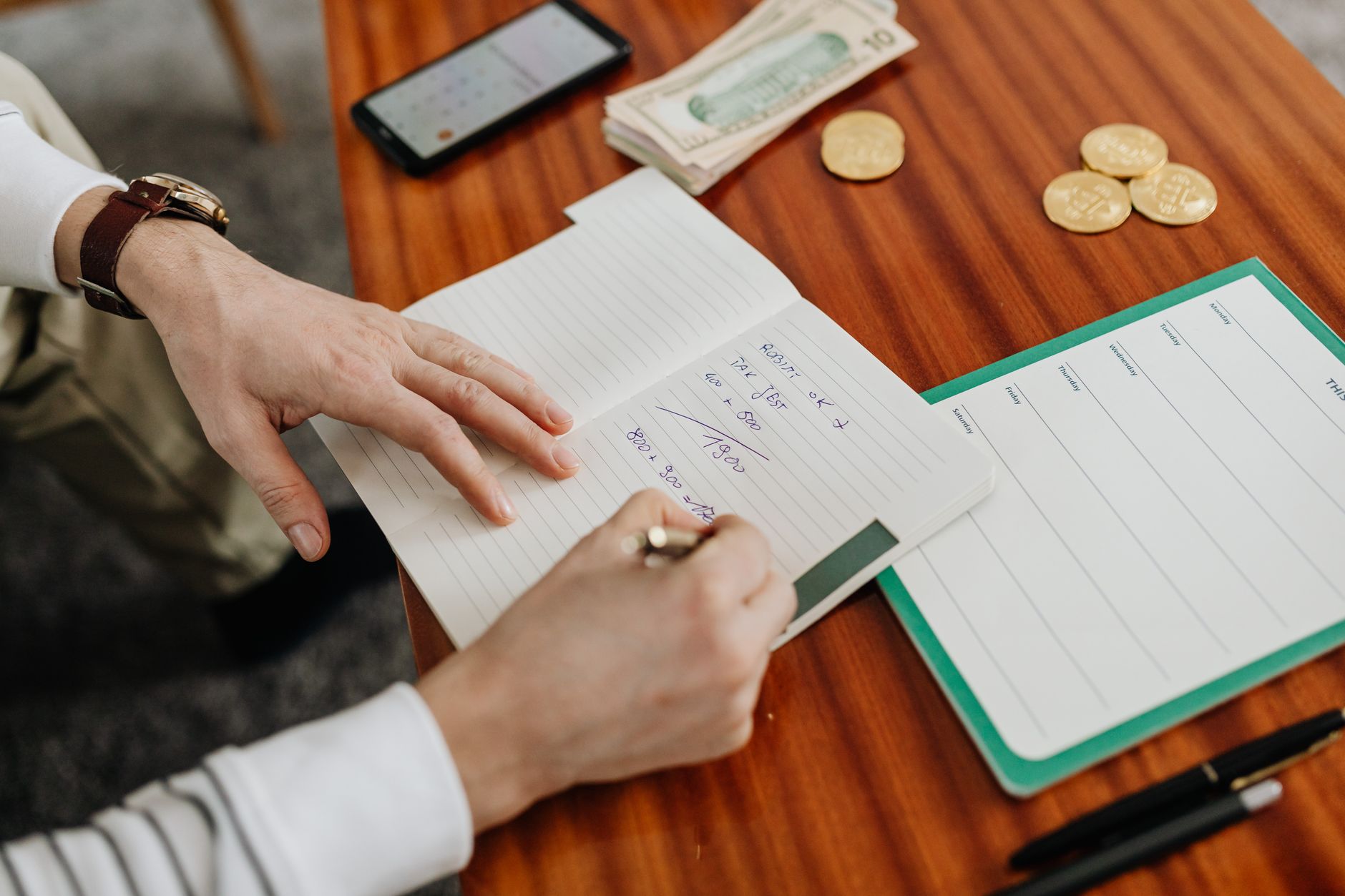Hand writing in a planner beside coins and a phone